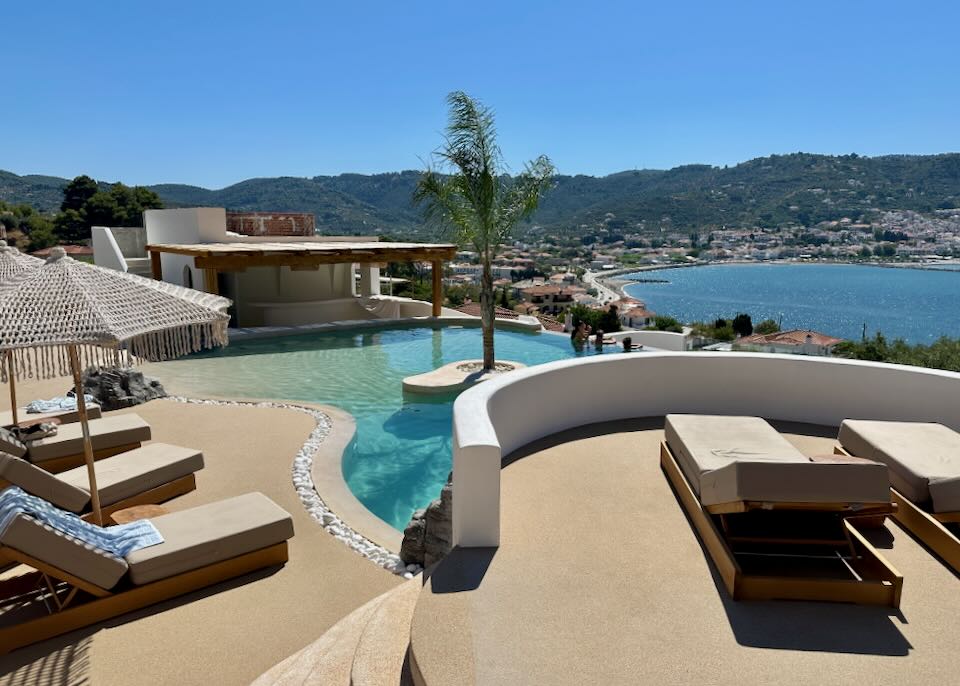 Hotel pool deck with infinity pool and shaded sun loungers, overlooking a Greek seaside village