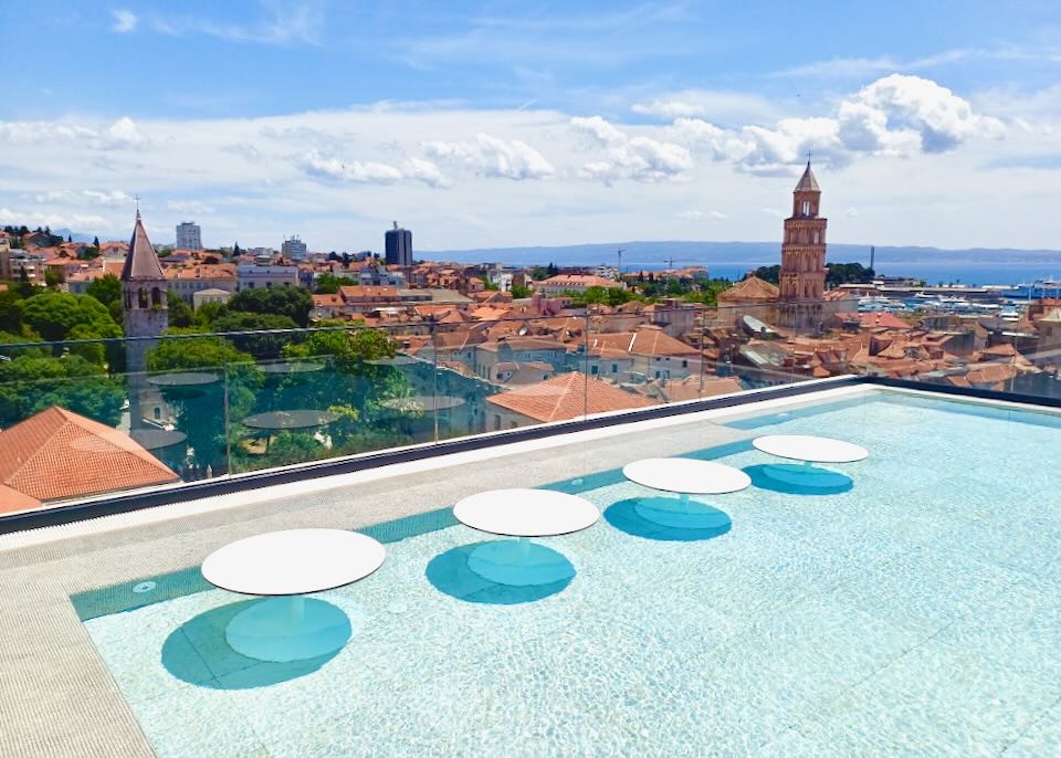 A pool with tables sits on a roof overlooking an old town.