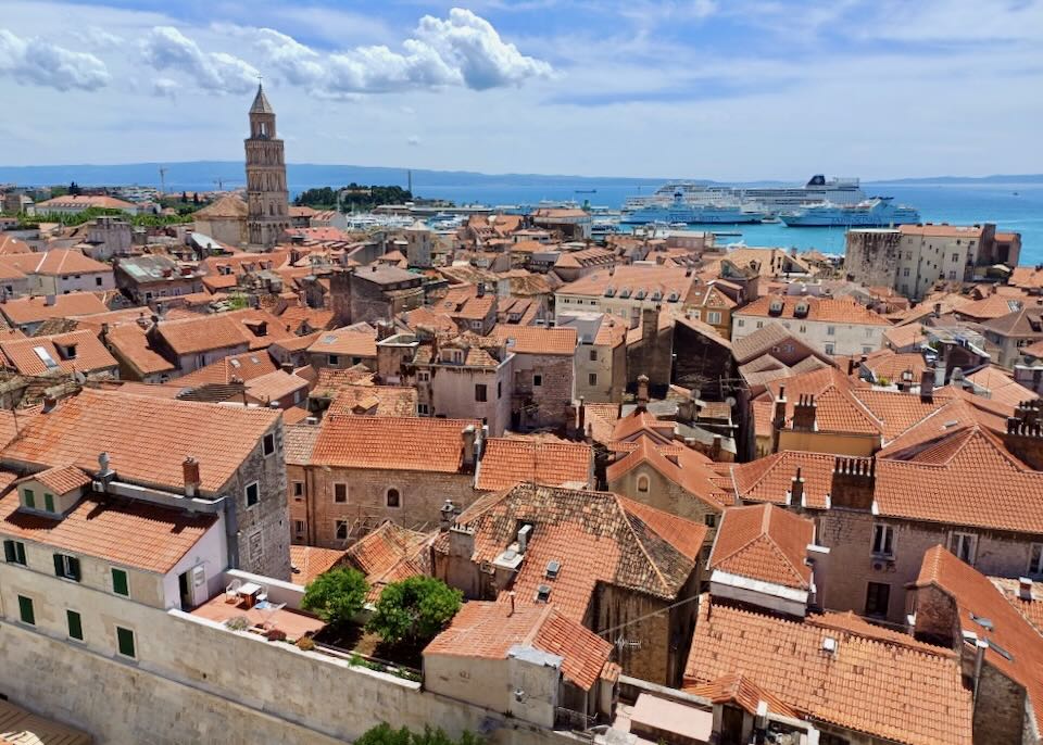 A view of buildings with terracotta roofs and a church tower, next to a port and blue water.