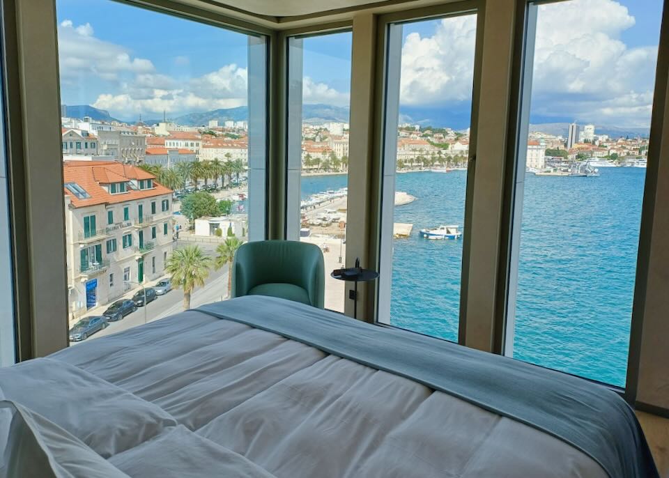 A bed in a hotel sits next to floor-to-ceiling window and a view of the waterfront.