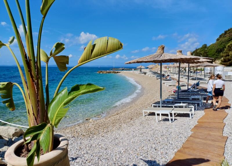 People walk on wood planks across sun-warmed stones to lounge chairs on a rocky beach.