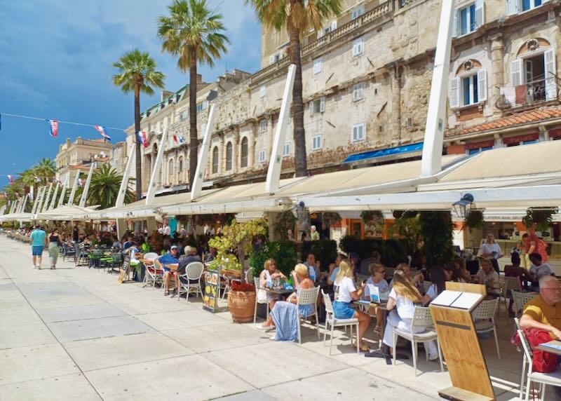 People site under awnings along a waterfront promenade.