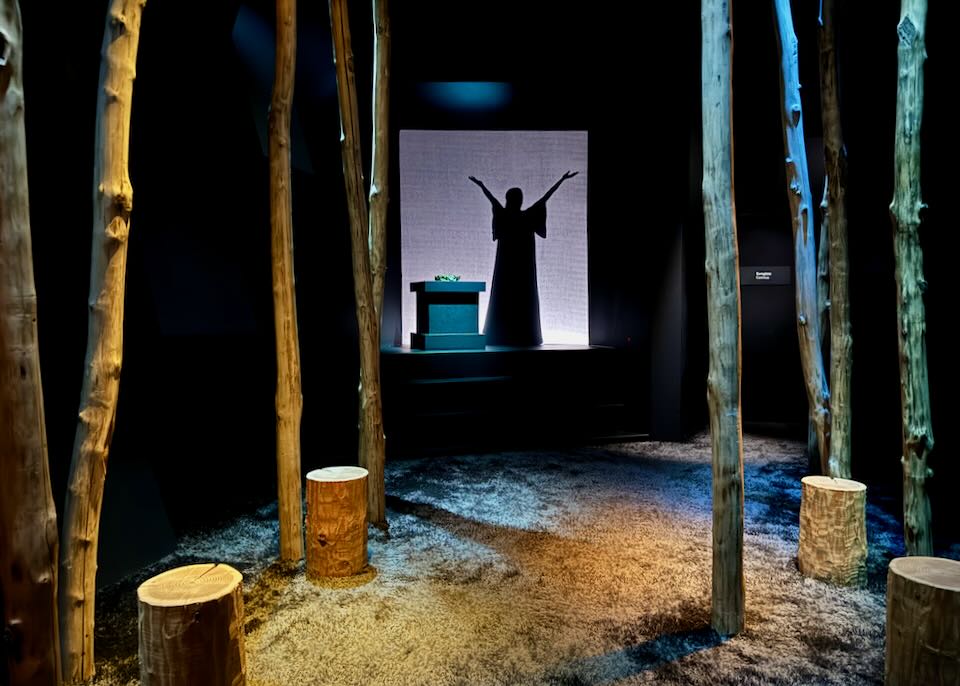 A silhouette of the opera singer Maria Callas stands on a stage in a room full of tree trunks, simulating a forest.