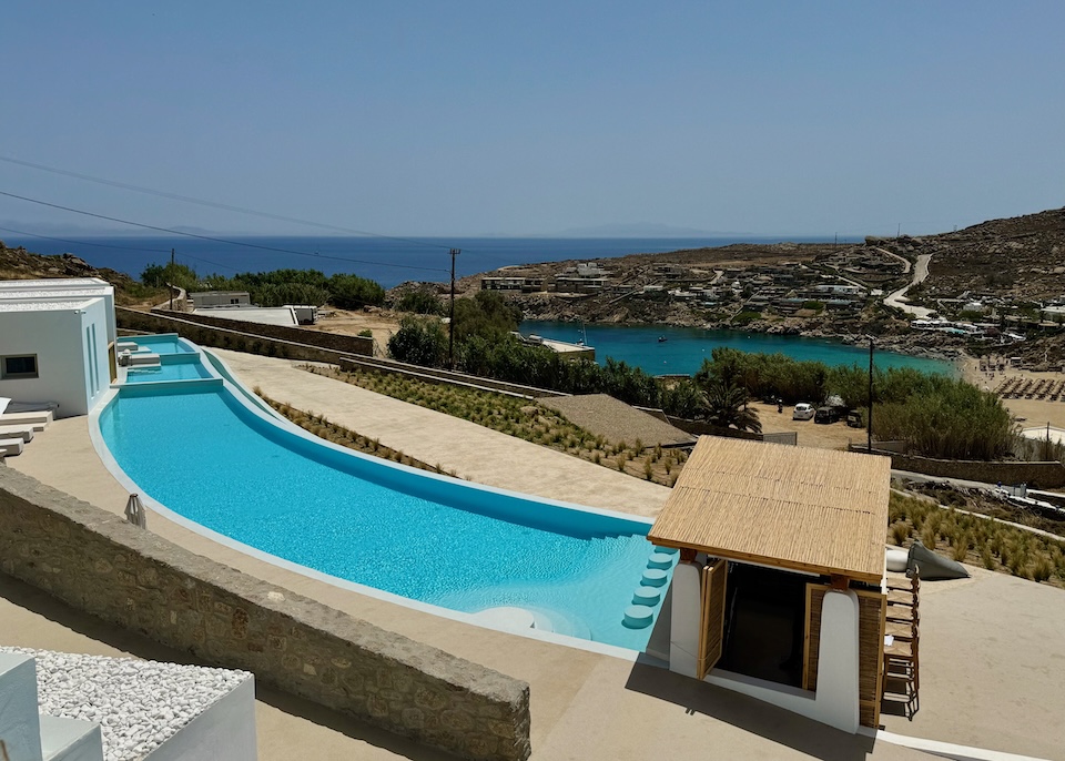 A curving infinity pool with a swim up bar at the near end a view toward the beach at Mykonos Flow near Super Paradise Beach.