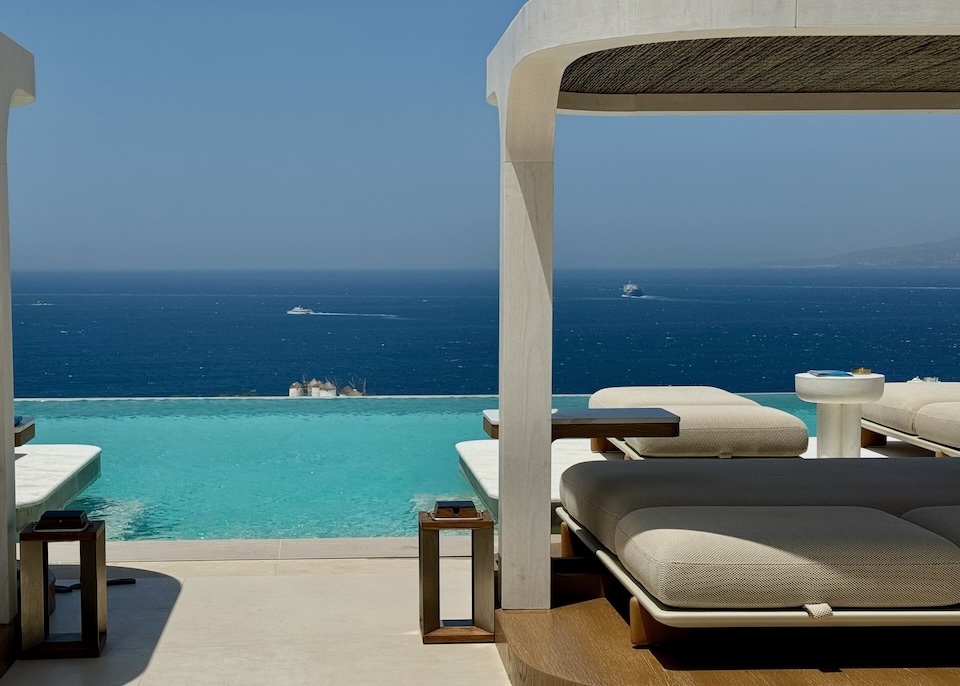 A contemporary poolside cabana behind an infinity pool with a view toward the Little Venice windmills and sea at Lovia in Mykonos Town.
