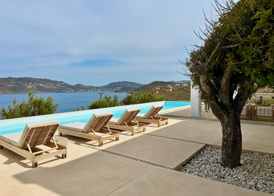A row of sunbeds behind an infinity pool on a terrace with a tree and sea view at the Margi Villa in Agios Sostis, Mykonos.