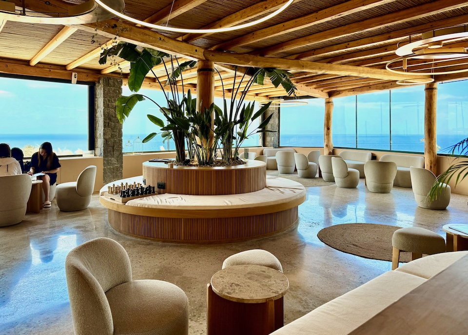 Contemporary lobby with a round sofa with a planter in the center, circle-shaped overhead lights, an exposed wooden ceiling, and a wraparound wall of windows with a view to the sea at Andronis Arcadia in Oia.