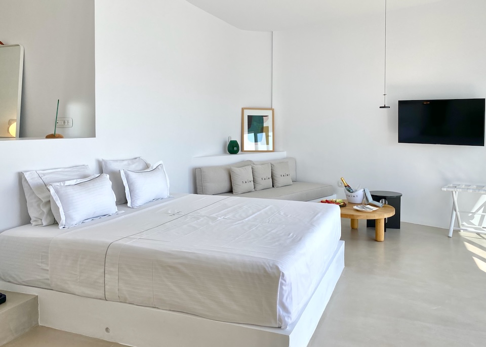 A whitewashed guest room with a king-size bed, sofa, and welcome bottle of wine and fruit platter at Saint in Oia.