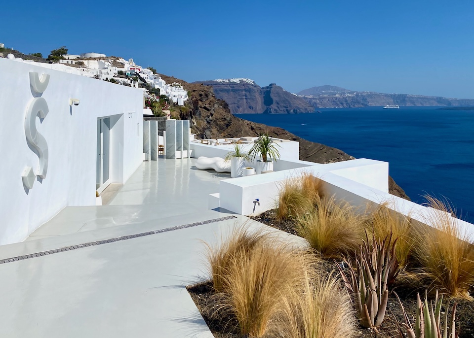 A building with a whitewashed exterior and a giant S on the side perched above the caldera at Saint hotel in Oia.