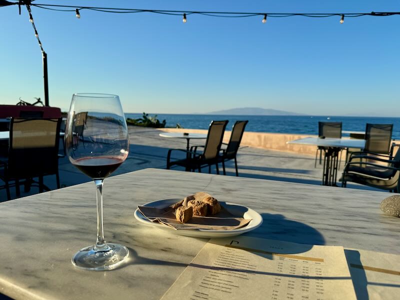 A glass of wine and plate of dried bread on a table looking out over the sea at sunset.