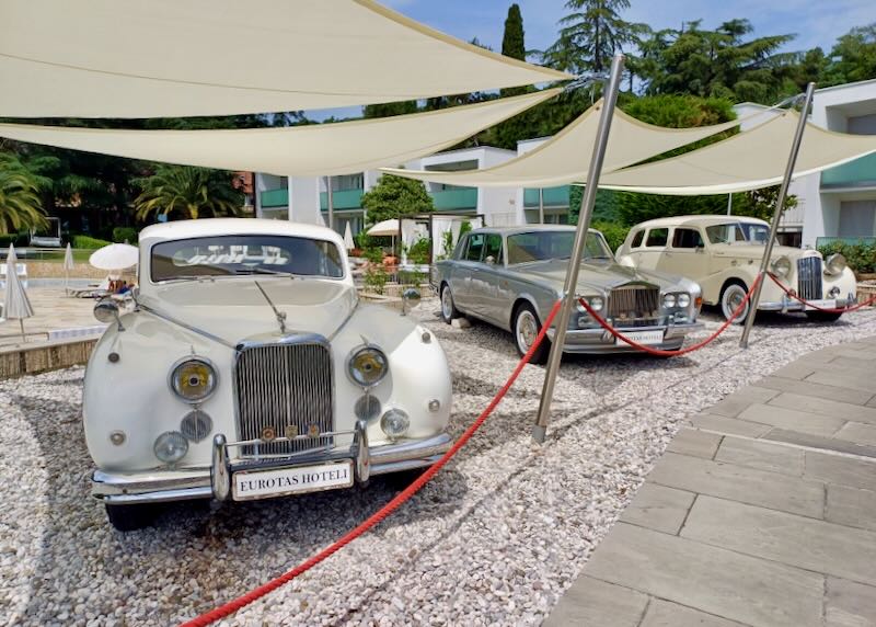 Three vintage cars sit under canvas canopies and behind red ropes.