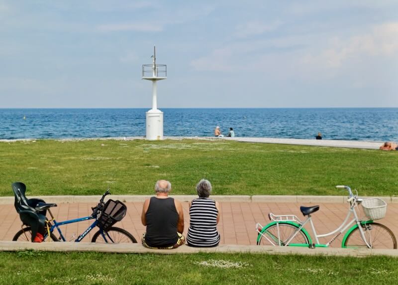A couple sits on a stone retaining wall, bikes leaning against it, as they watch the ocean.