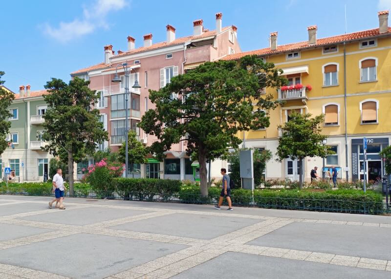 People walk on a large sidewalk past green trees and pink, pale green, and gold painted buildings.