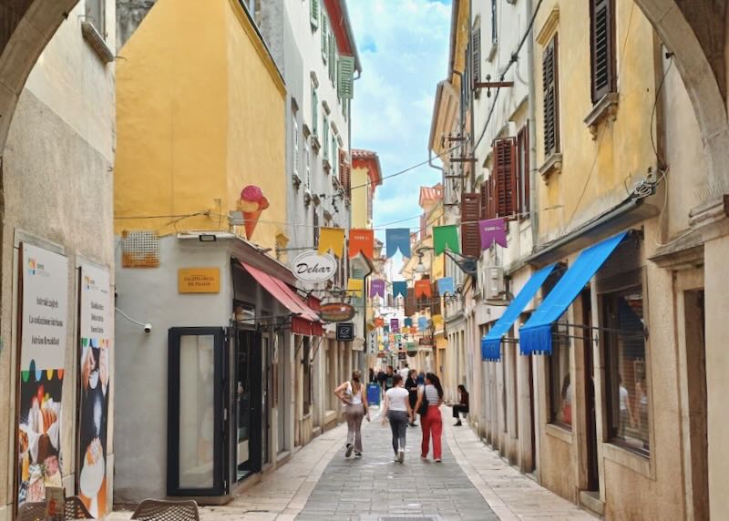 Three women walk down a stone path by colorful buildings and shops.
