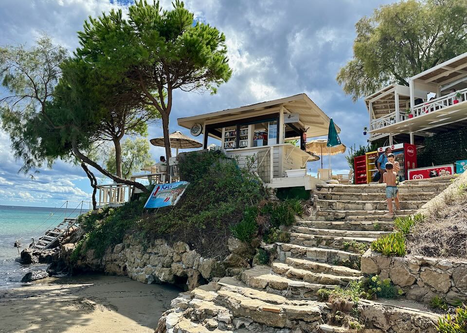 Stone steps leading up to a small cafe on a beach, shaded by a tree.