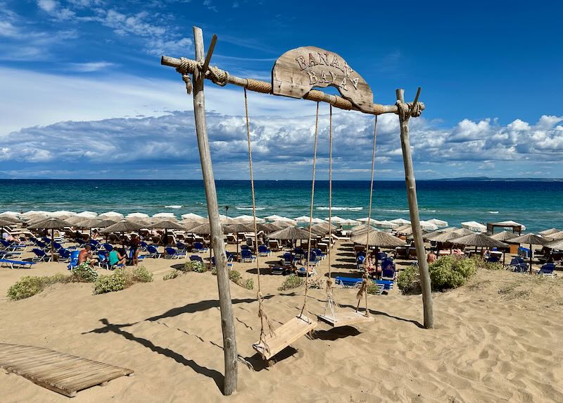 entrance to a golden sandy beach with wooden swings advertising the name of a beach bar