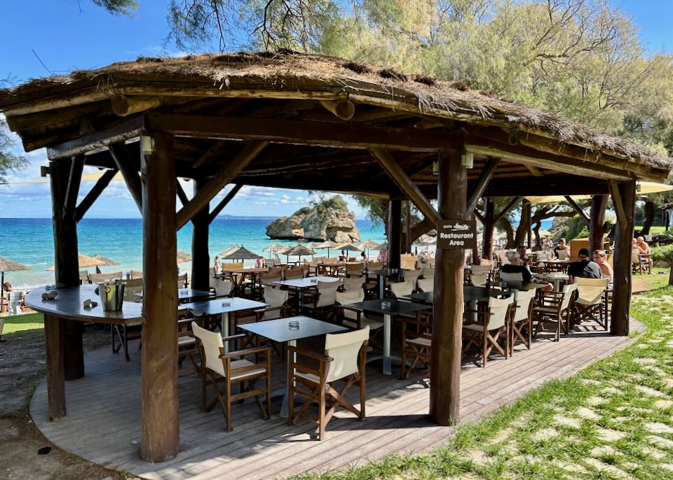 Open air, covered restaurant tables overlooking a beach in Greece.