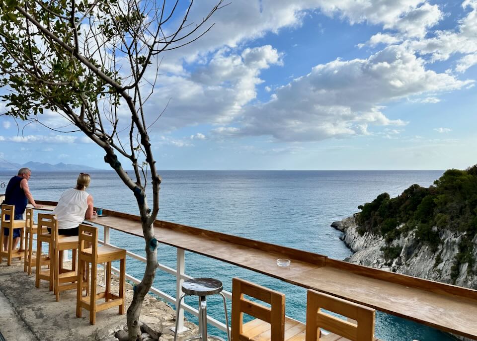 Two people sit at bar seating overlooking a scenic beach in Greece