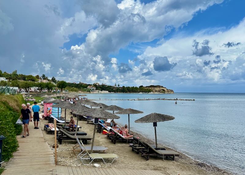 People walk on a wooden boardwalk along a narrow beach lined with sun umbrellas