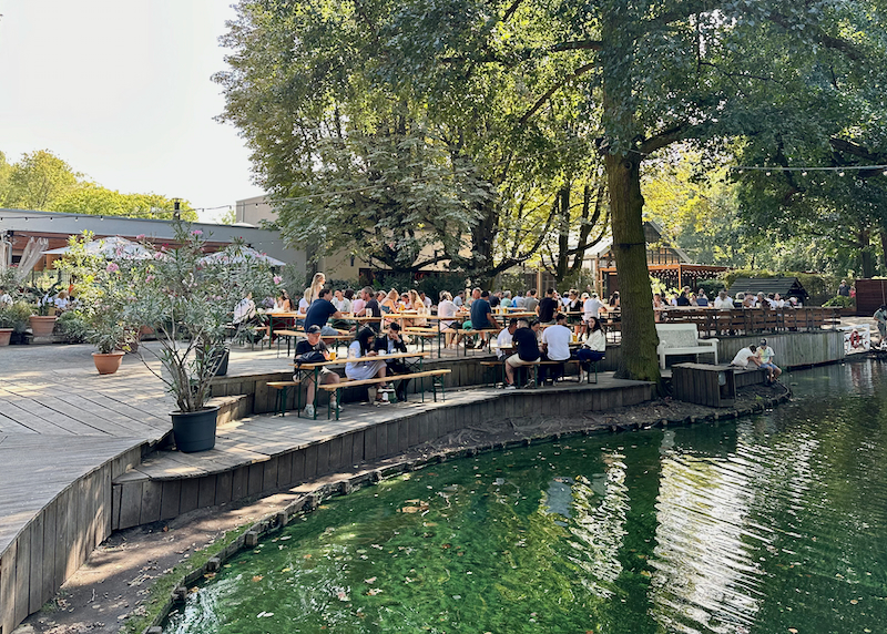 People dine at picnic tables beside a tree-lined lake