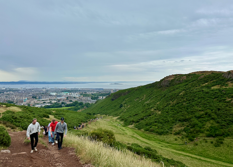 People hike on a large green, grassy hill rising above a city. 