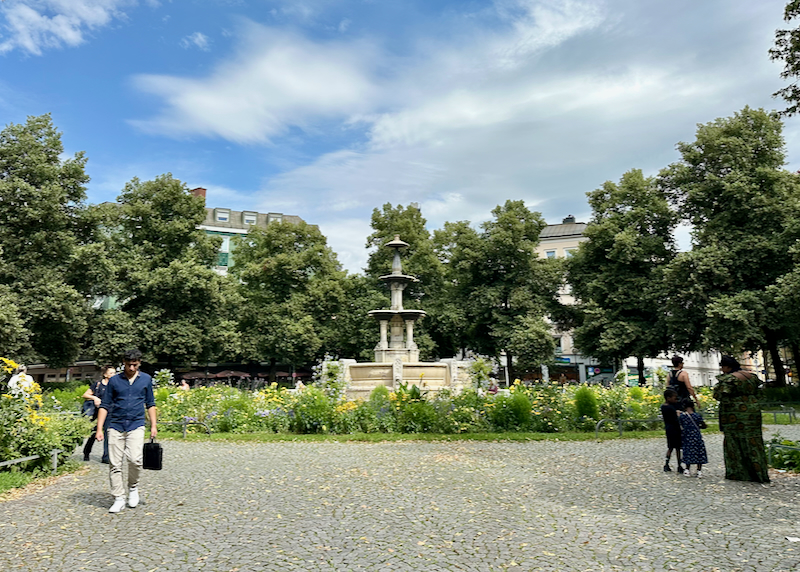 A man walks by with a briefcase and a older woman helps a young child by a park fountain