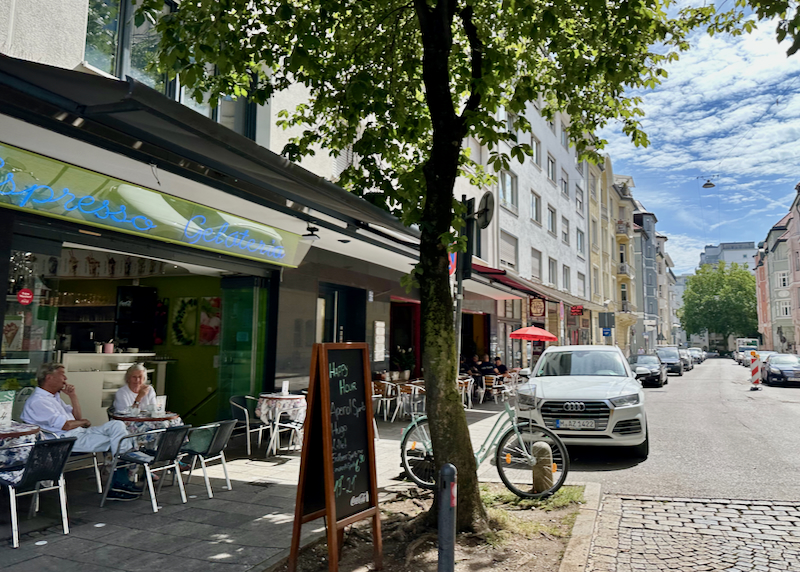 People dine at sidewalk cafe tables on a leafy street in Munich