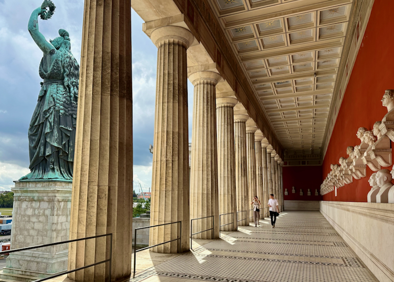 A man and a woman walk past down a colonnade displaying busts of famous Bavarians
