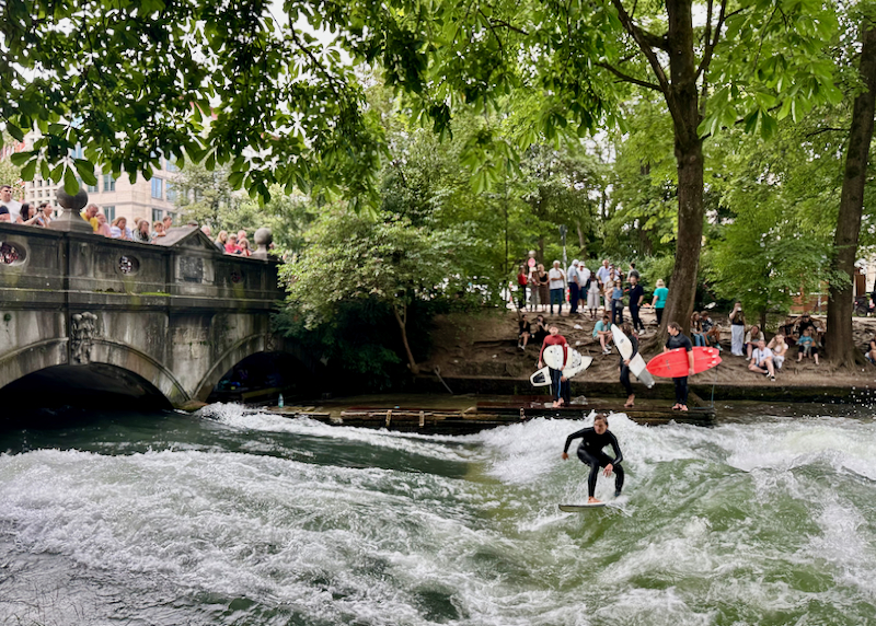 A woman surfs a wave in an urban river, while onlookers watch from a bridge above