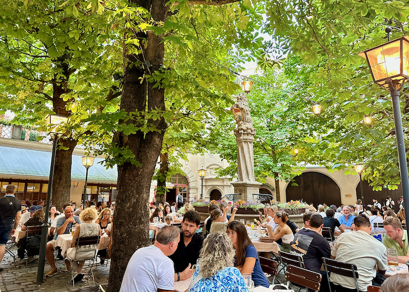 A crowded outdoor beer garden, shaded by trees and with a statue at the center