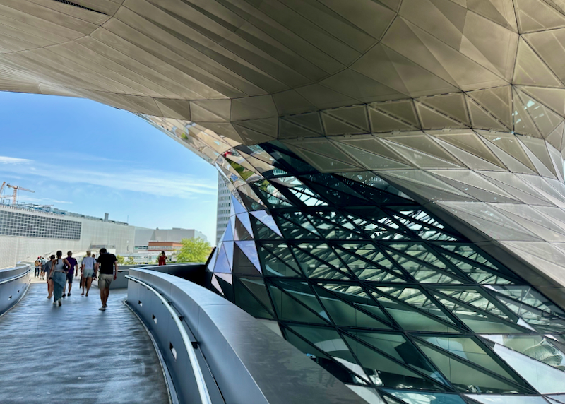 People walk along a sidewalk past a curved glass arch