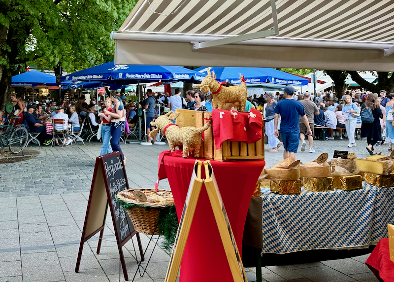 Tables with cheese displayed at an outdoor market, as pedestrians pass by.
