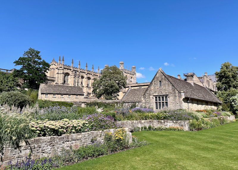View of the gothic spires of Oxford's Christ Church College from a green lawn bordered by a flowering herb garden