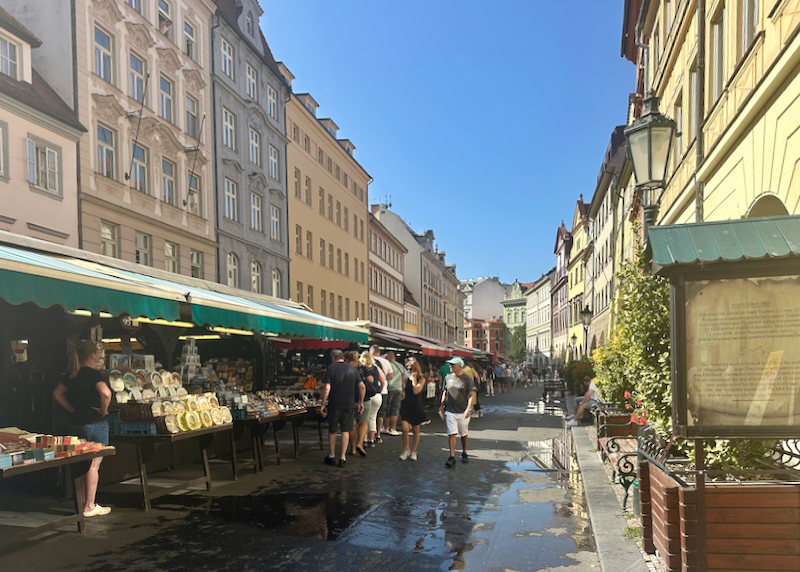 People walk by market stands on a street lined with colorful buildings.