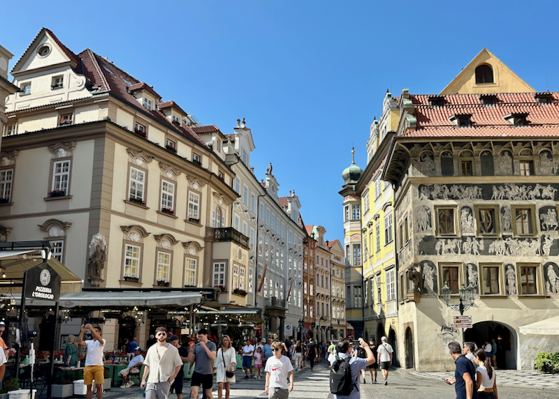 pedestrians walk through a square lined with decorated Czech buildings