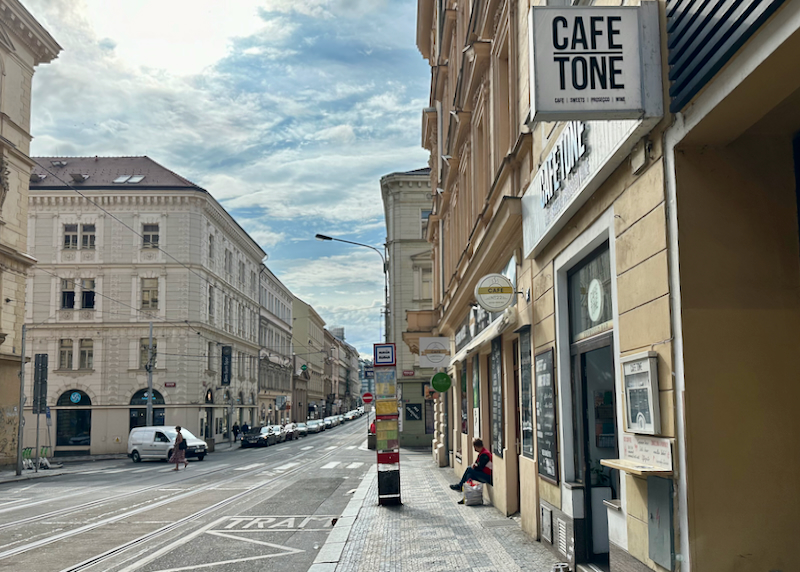 A man sits on a stoop of a small shop on a Czech street lined with cafes and small stores