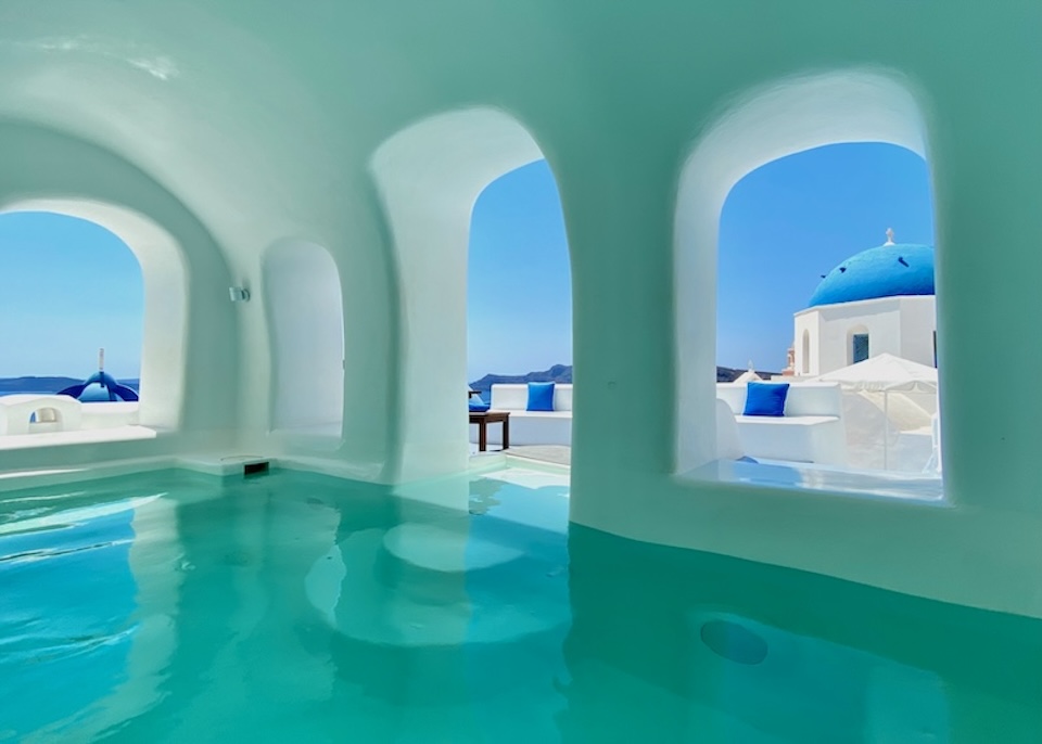 A pool inside a manmade cave with glassless windows and views of Santorini's blue domes and caldera at Oia Spirit Boutique Hotel.