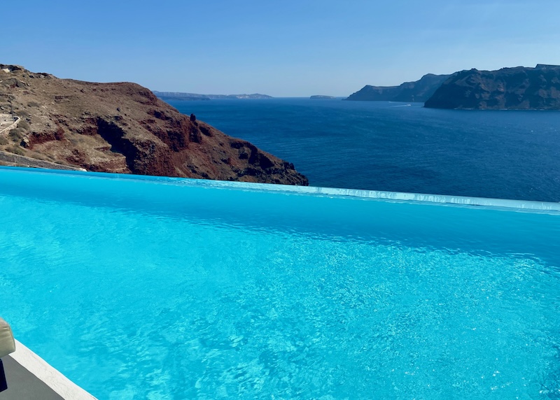 Infinity pool over the sea with a view toward Thirassia Island and the switchback path down to Ammoudi Bay in Santorini.