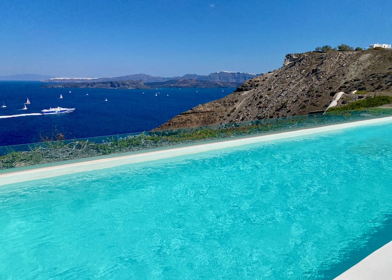 A long infinity pool above the caldera with views toward sailboats, the volcanoes, and Oia village in the distance at Cocomat hotel in Santorini.