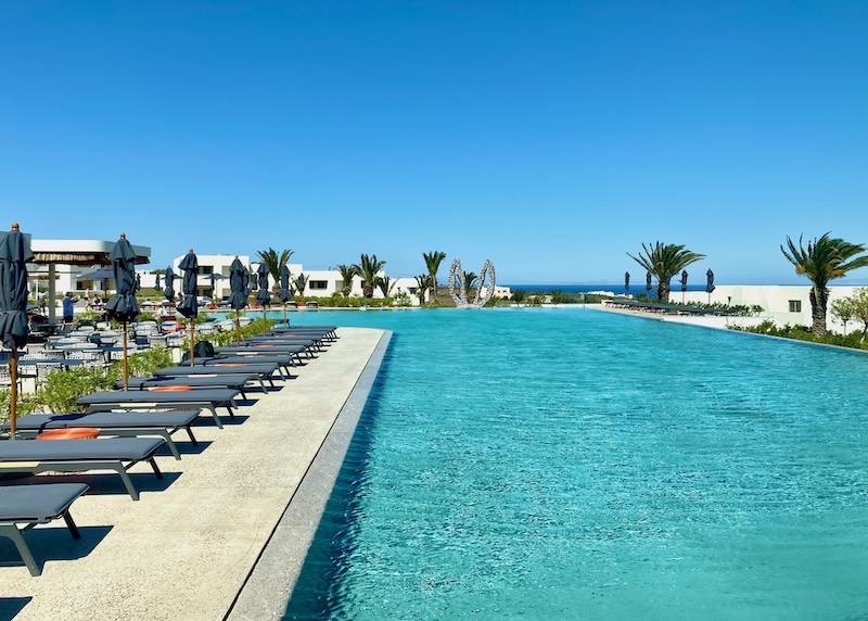 An L-shaped infnity pool lined with sunbeds and palm trees at Nous Resort in Santorini.