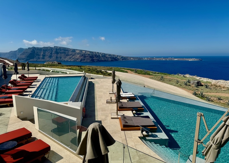 Two infinity pools lined with sunbeds and umbrellas with views over the sea toward Thirassia Island at Santo Mine in Santorini.