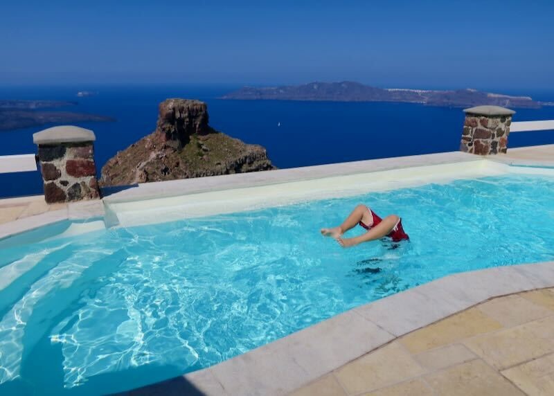 A child doing a handstand in an infinity pool overlooking the caldera, Skaros Rock, and Thirassia Island at Tholos Resort in Santorini.