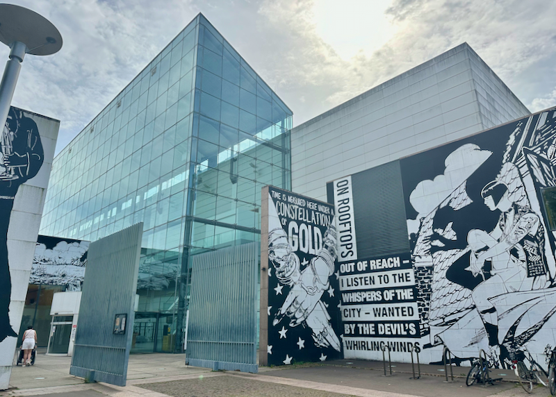 Angular exterior of a glass-and-steel museum building, with a black-and-white mural displayed in front