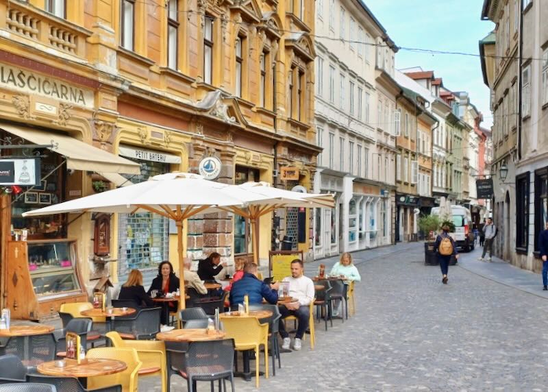 People sit at tables at a café on stony streets surrounded by tall historic buildings.
