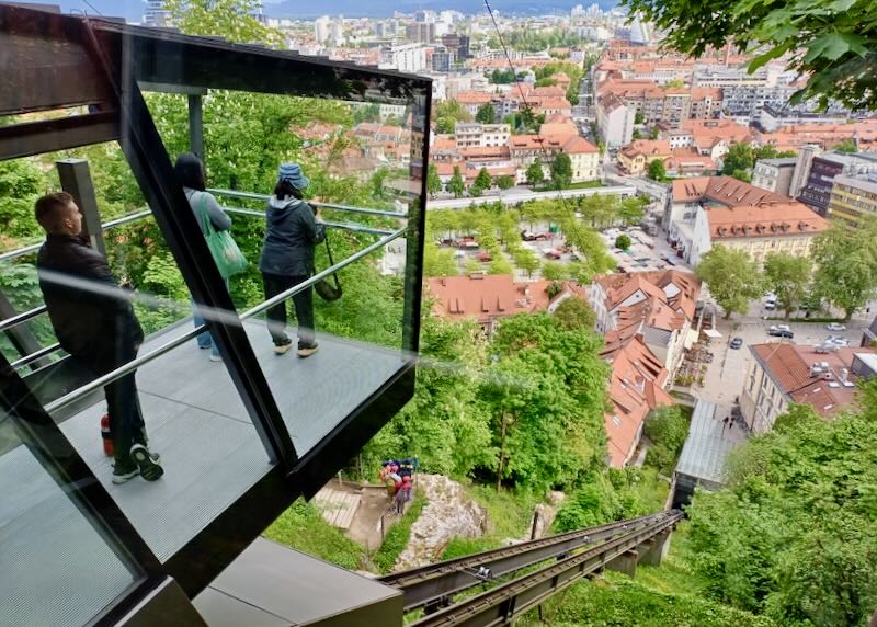 People look out a glass funicular to a city with orange tile roofs.