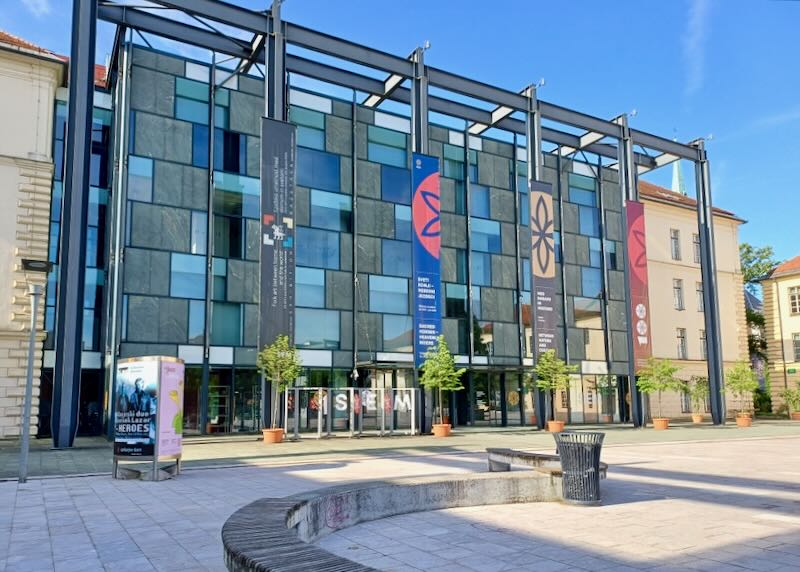 The exterior of a multi-story museum, has banners hanging on steal pillars.