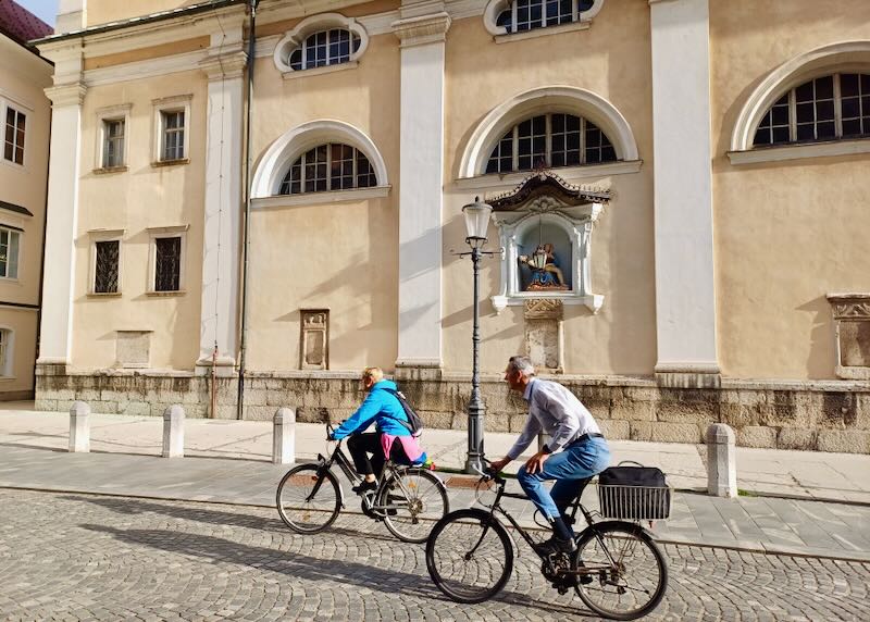 A couple rides bikes on stony roads by an old church.