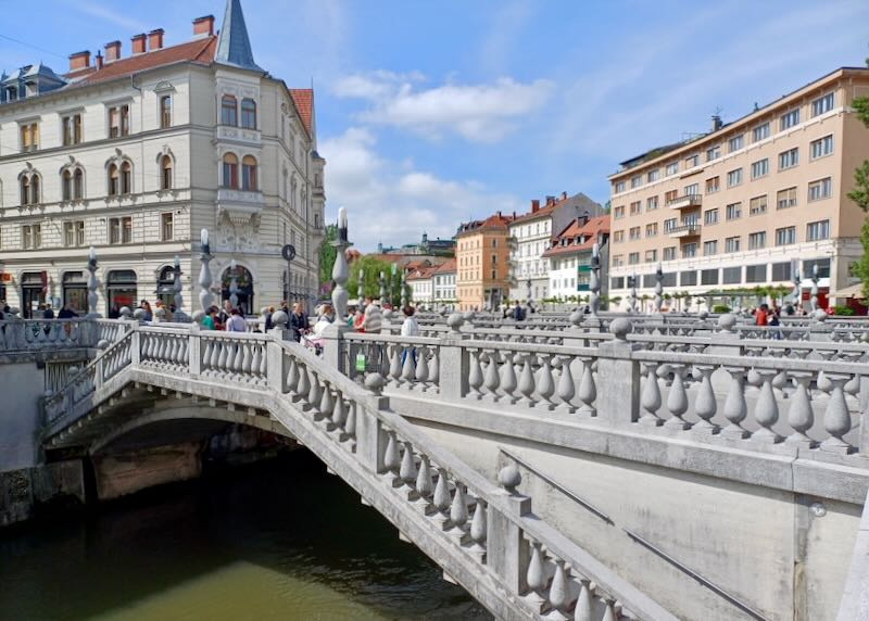 A stone bridge crosses a green river to tall old pastel-colored buildings.