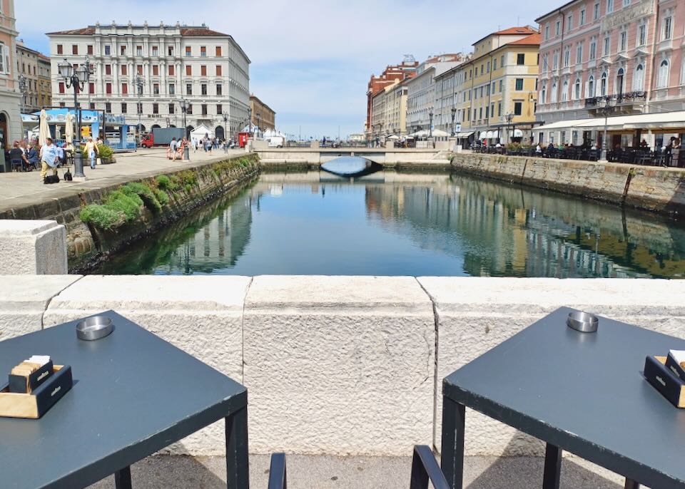 Two black tables sit by a canal with views of old pastel-painted buildings and the sea.