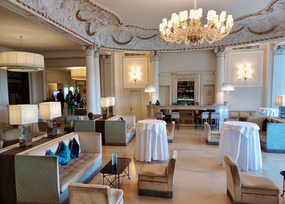 An ornate gold and white ceiling with a glass chandelier sits above a lobby in a hotel.